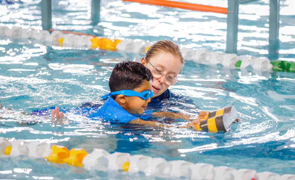 Photo of young boy swimming in the pool with a kickboard in hands, beside female swim teacher as she guides him through a swimming exercise.
